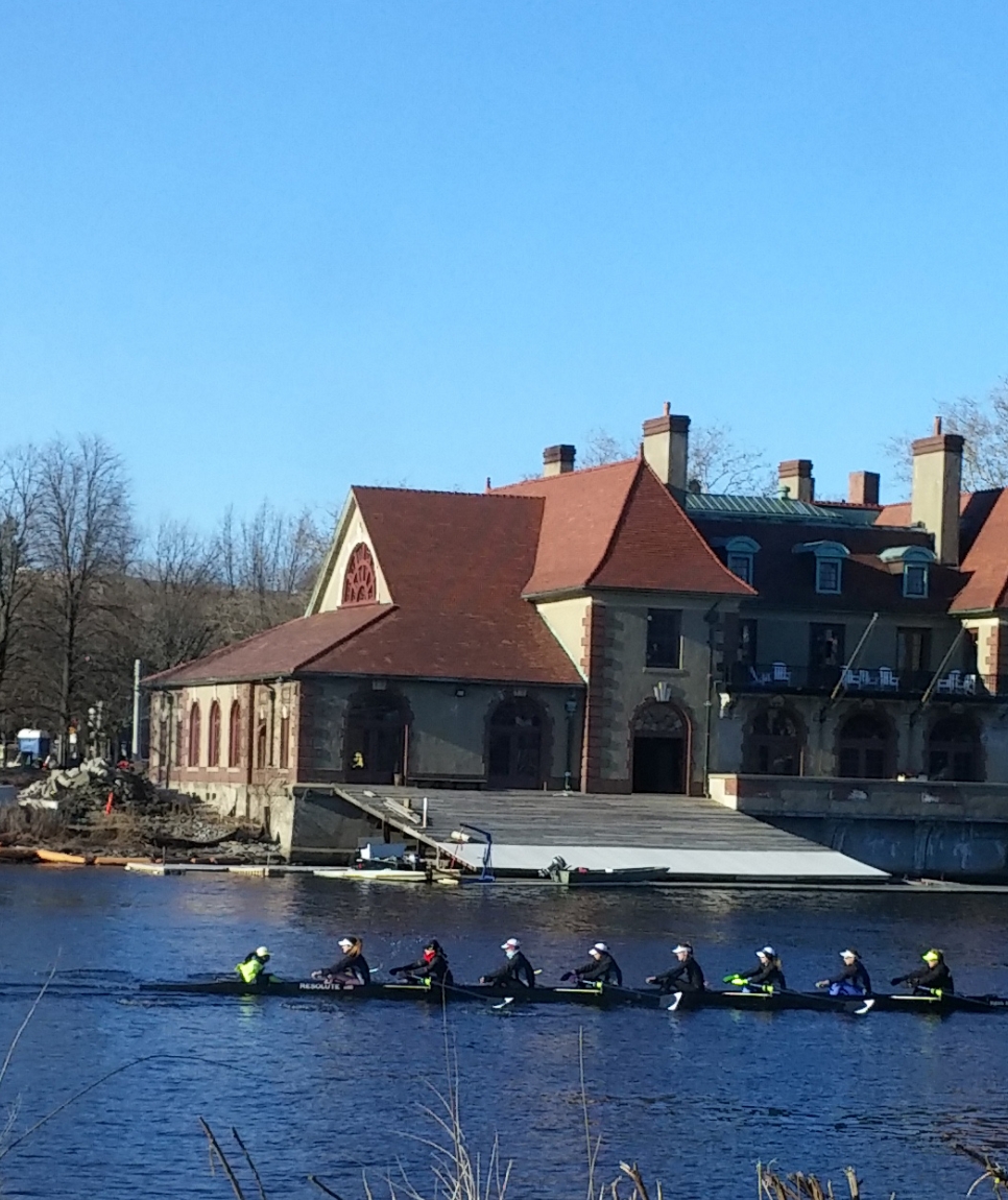 A rowing crew training on a river beside a campus boathouse